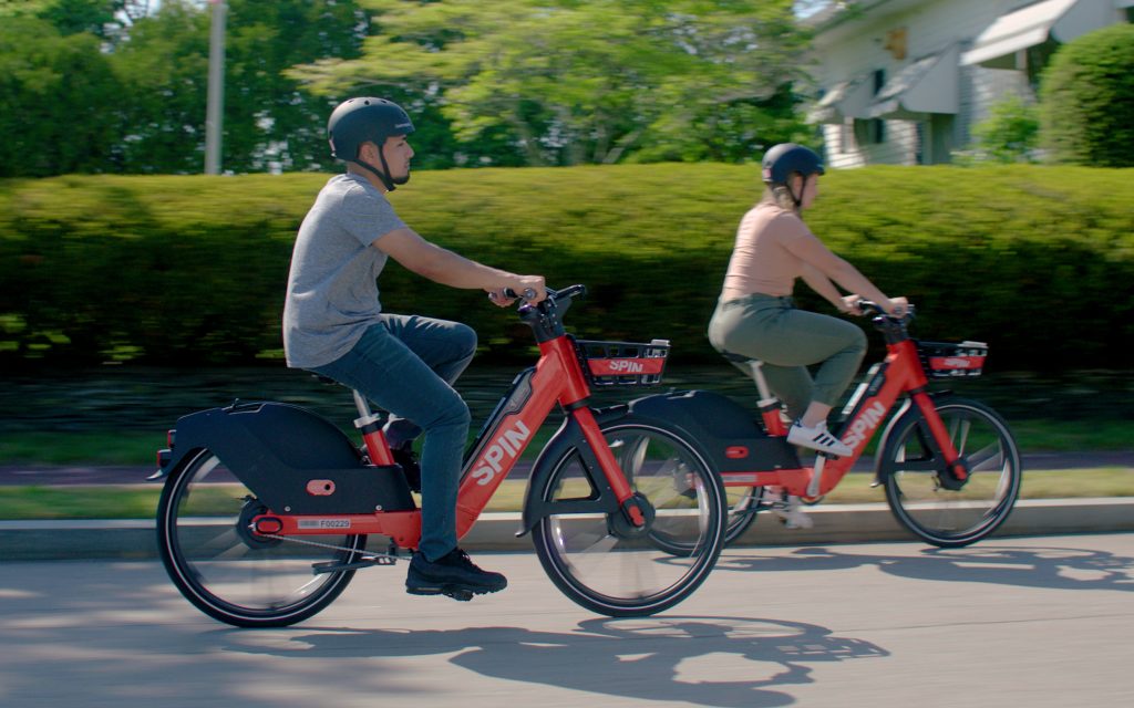 two individuals riding orange SPIN e-bikes in a neighborhood