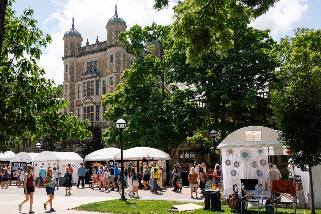 Crowds walk down South University Avenue past vendor tents under the lush green trees in front of the Law Quad during the 2024 Ann Arbor Art Fair.