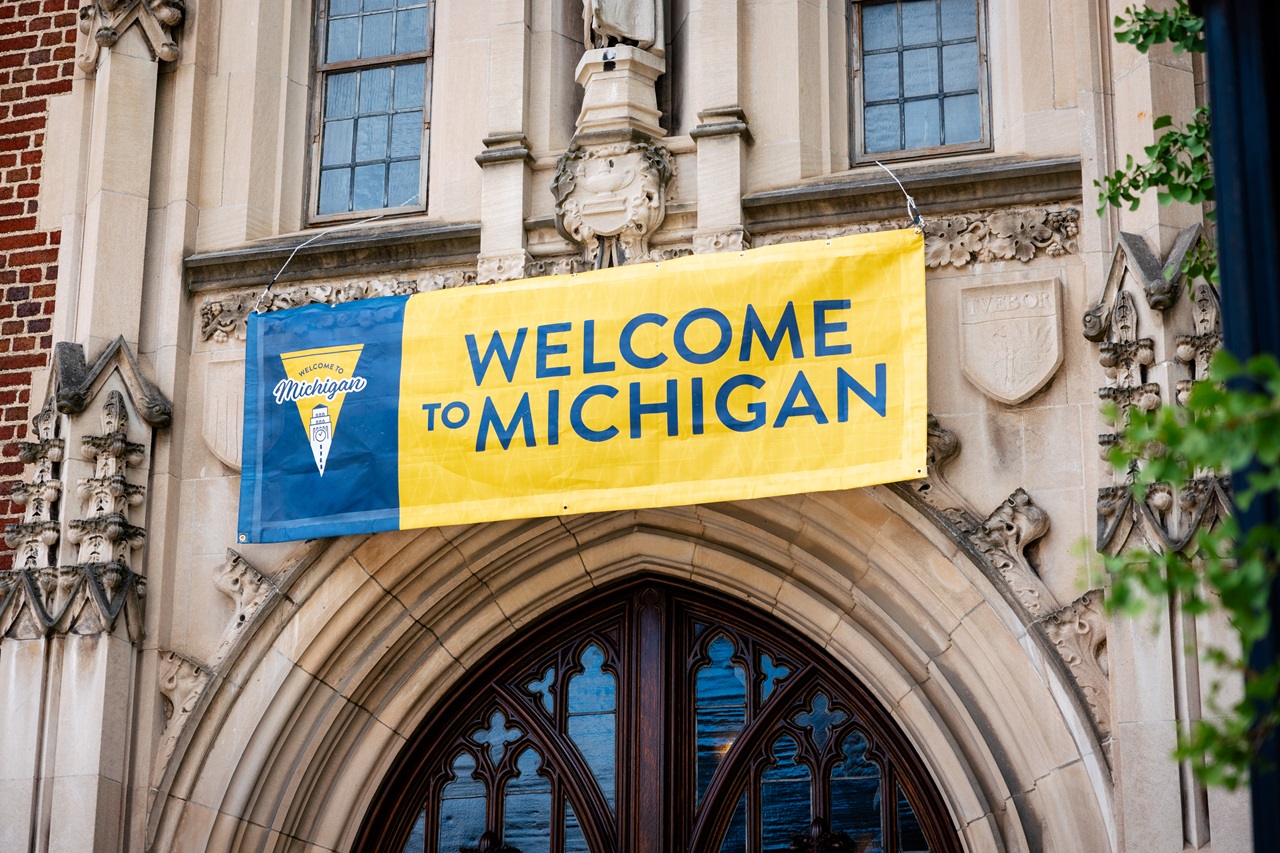 A blue and yellow banner with a pennant design and illustration of Burton tower and the text “Welcome to Michigan” hangs above the doorway to Martha Cook ahead of 2024 Fall Move-In.