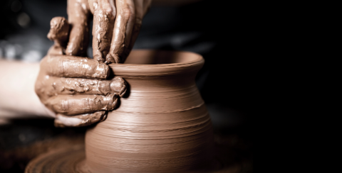 Close-up of hands shaping a clay pot on a pottery wheel against a dark background.